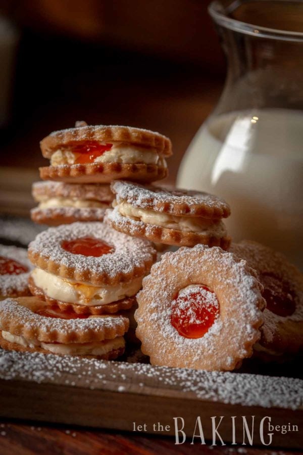 Linzer cookies stacked on a plate dusted with powdered sugar, next to a pitcher of milk