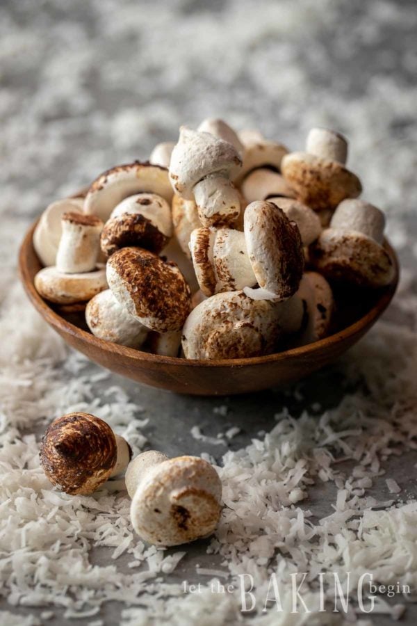 Meringue mushrooms piled in a bowl that is sitting amongst scattered shaved coconut.