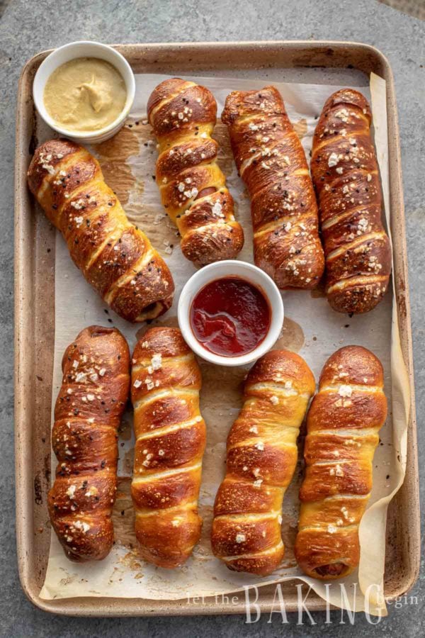 Pretzel dogs on a baking sheet with mustard and catchup in small round mini bowls.