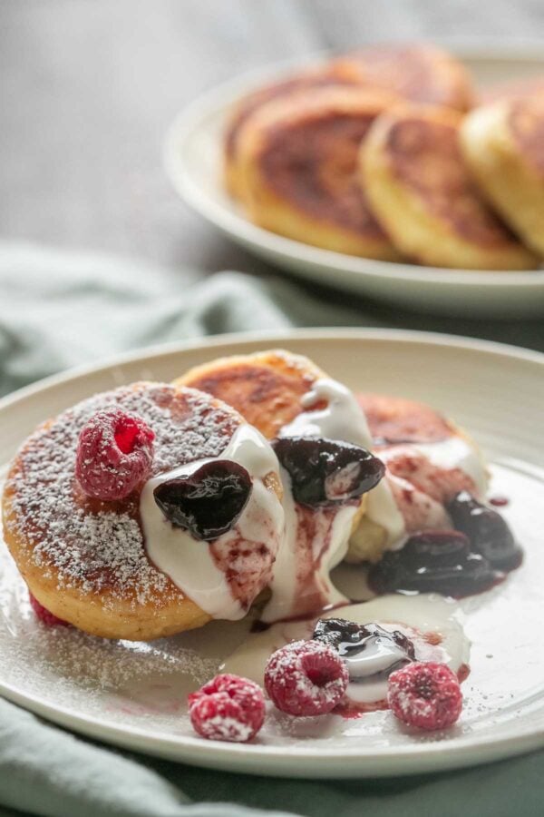 Syrniki pancakes on a plate topped with powdered sugar, sour cream, and jam.