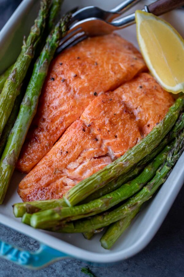 Two air fried salmon fillets surrounded by asparagus and a lemon wedge in a ceramic baking dish.