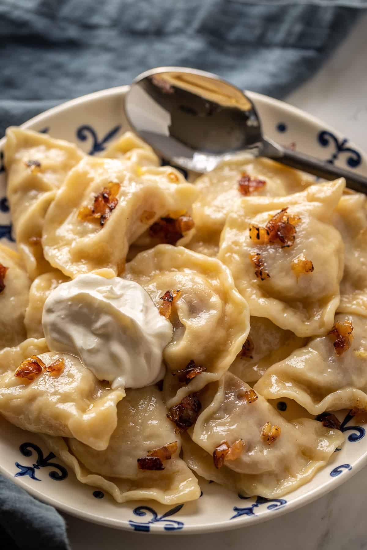 A plate full of potato perogies, sprinkled with caramelized onions and a dollop of sour cream, with a blue napkin in the background.