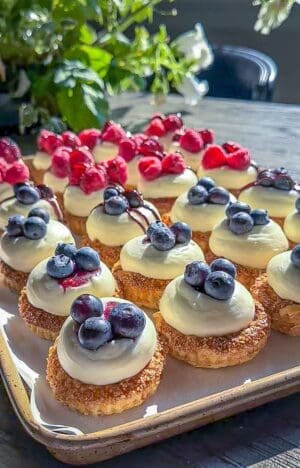 Puff pastry tarts filled with cheesecake filling and a berry center sitting on a baking sheet with some garnished with blueberries and others with raspberries.