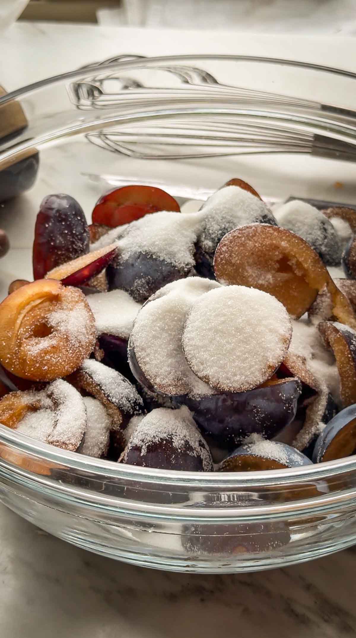 Halved Italian prune plums tossed with sugar in a glass bowl, macerating for a plum galette