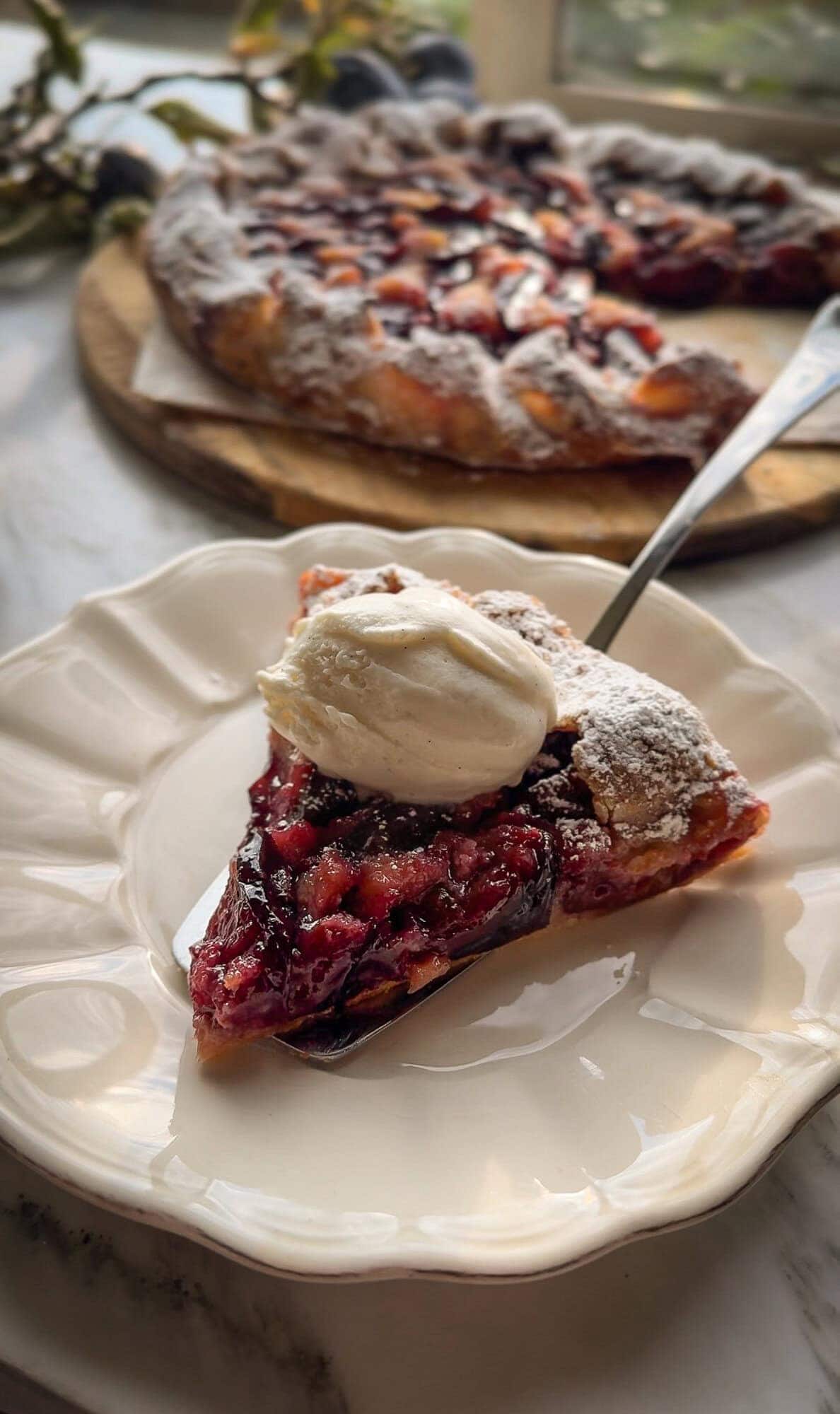 A slice of plum galette with almond cream topped with vanilla ice cream, the whole galette behind