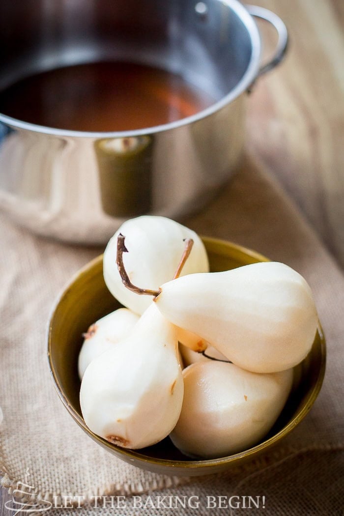 Peeled Bosc pears with stems on, next to a pot of amaretto poaching liquid